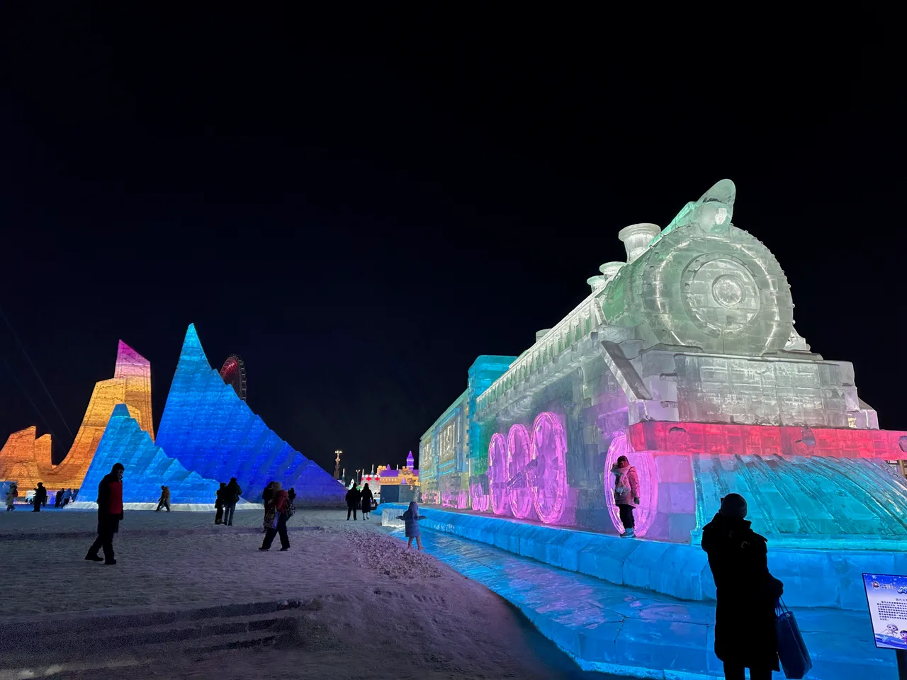 A large ice sculpture of a steam locomotive lit pink and blue at night, with visitors posing beside its wheels