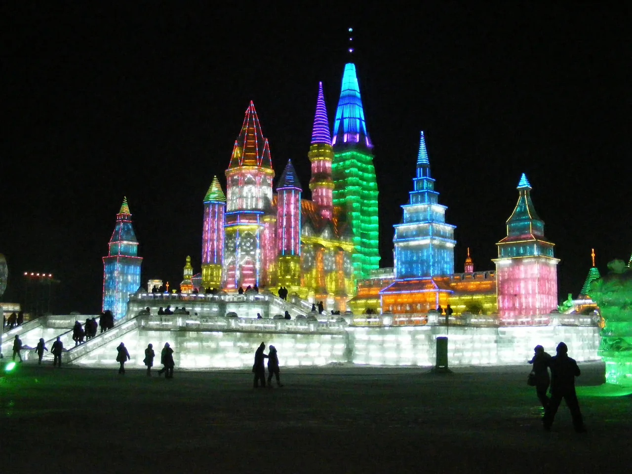 Ice buildings lit in pink, blue and green at night, with small figures of visitors walking along the base