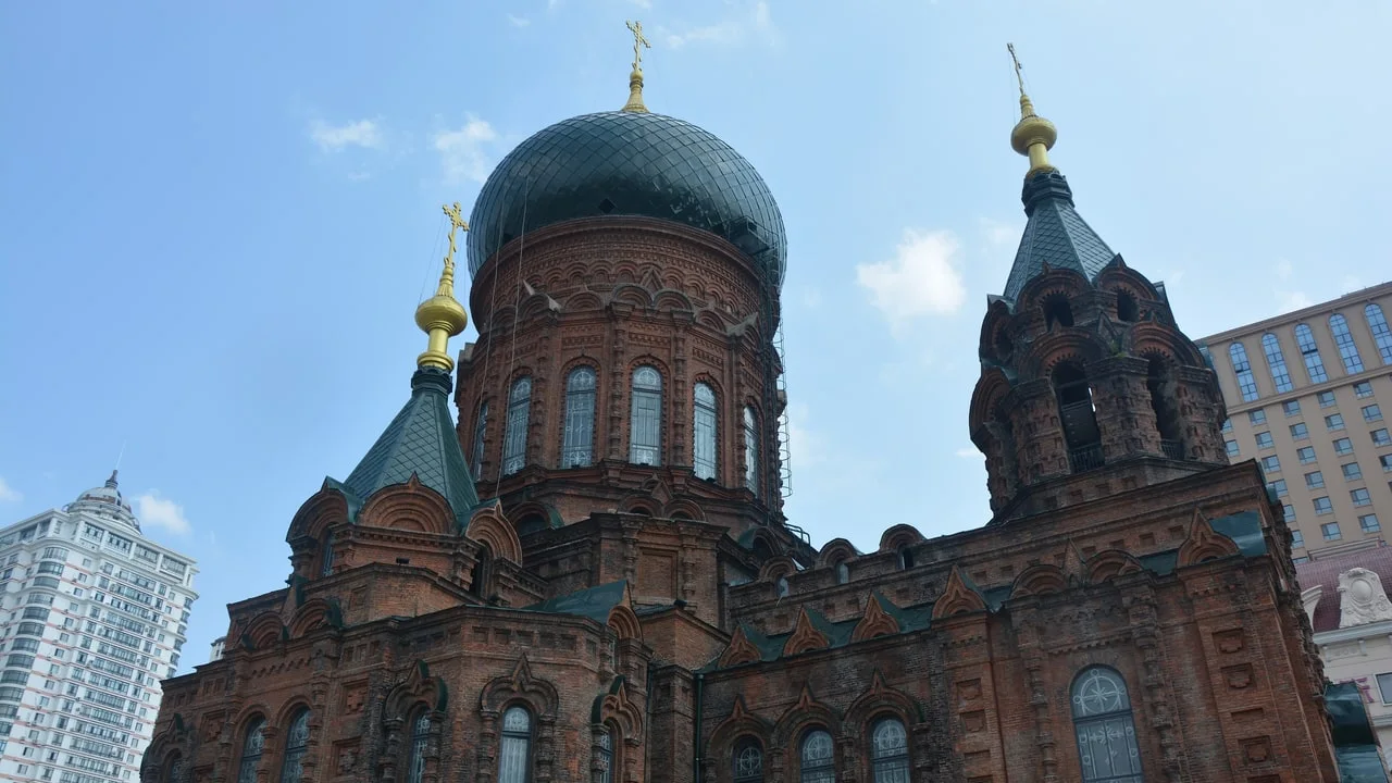 The green onion domes and red-brick towers of St Sophia Cathedral in Harbin against a blue sky, with modern tower blocks on either side