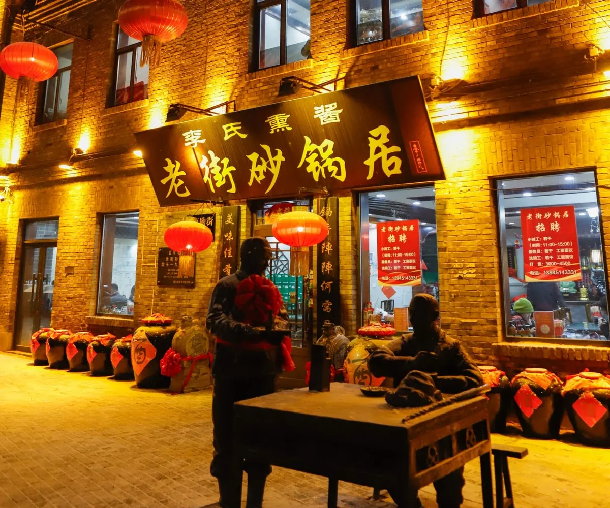 A brick restaurant front hung with red lanterns and a carved signboard at night, with bronze figures of a man standing and a man sitting at a table outside