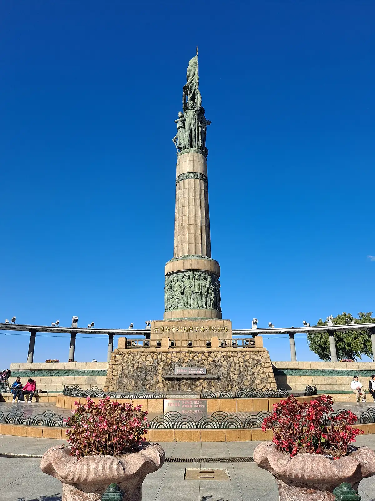A tall stone column topped by a group of bronze figures, rising from a granite base above a curved colonnade, under a deep blue sky