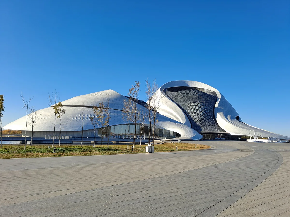 The white curved shell of the Harbin Grand Theatre, with a spiral of glass and a wide stone plaza in front, under a cloudless sky