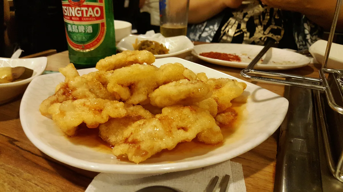 A white plate of golden battered pork slices glazed in a glossy sweet-and-sour sauce, with a bottle of beer and side dishes on the table