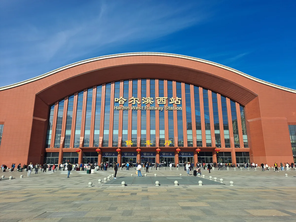 The long red-brick front of Harbin West railway station with a curved roof and tall glass arch, a wide paved forecourt and travellers in the distance
