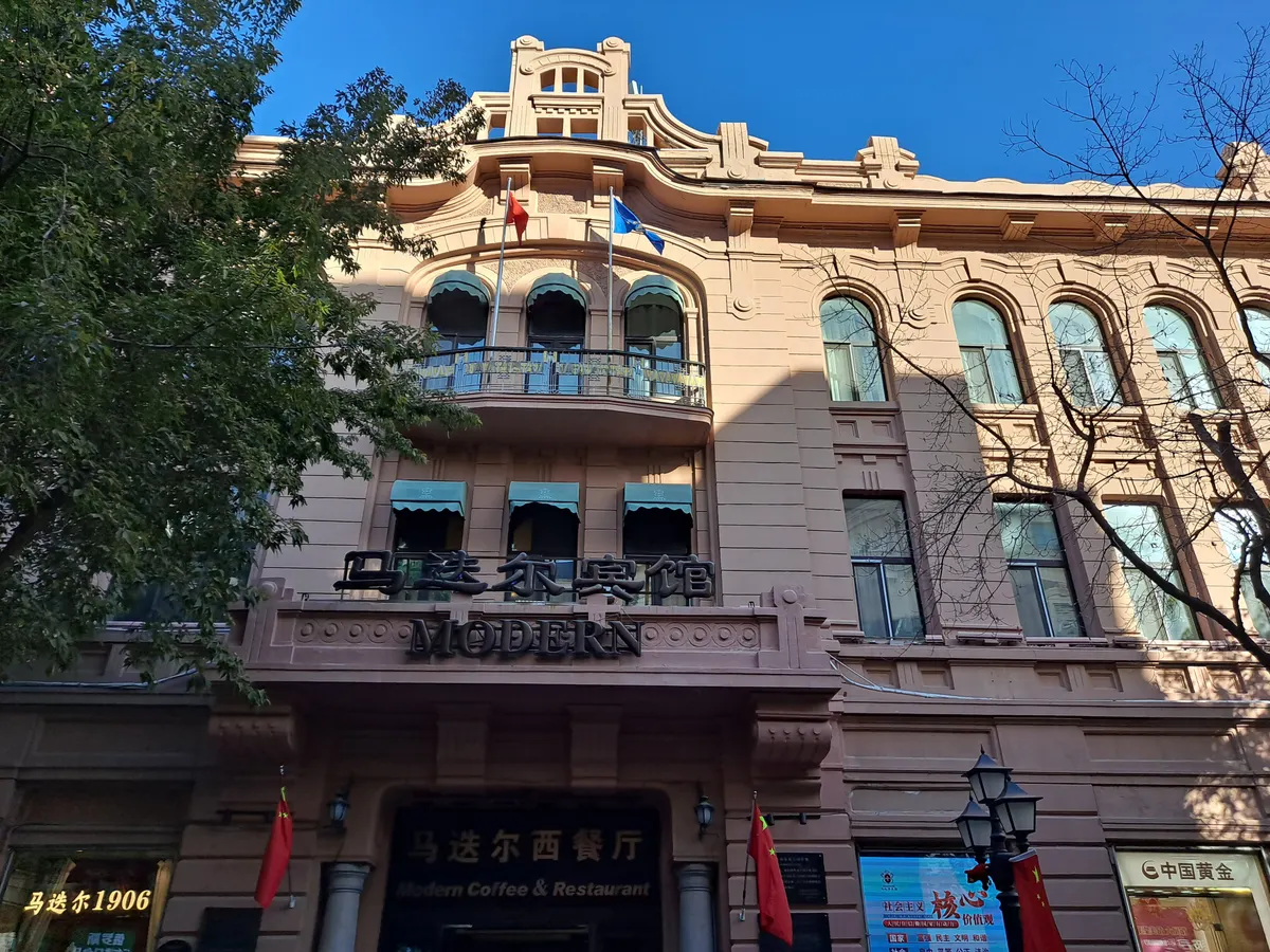 The pink facade of the Modern Hotel on Central Street, with its Chinese and Latin name above the restaurant entrance, flags on the balcony and a tree in autumn colour