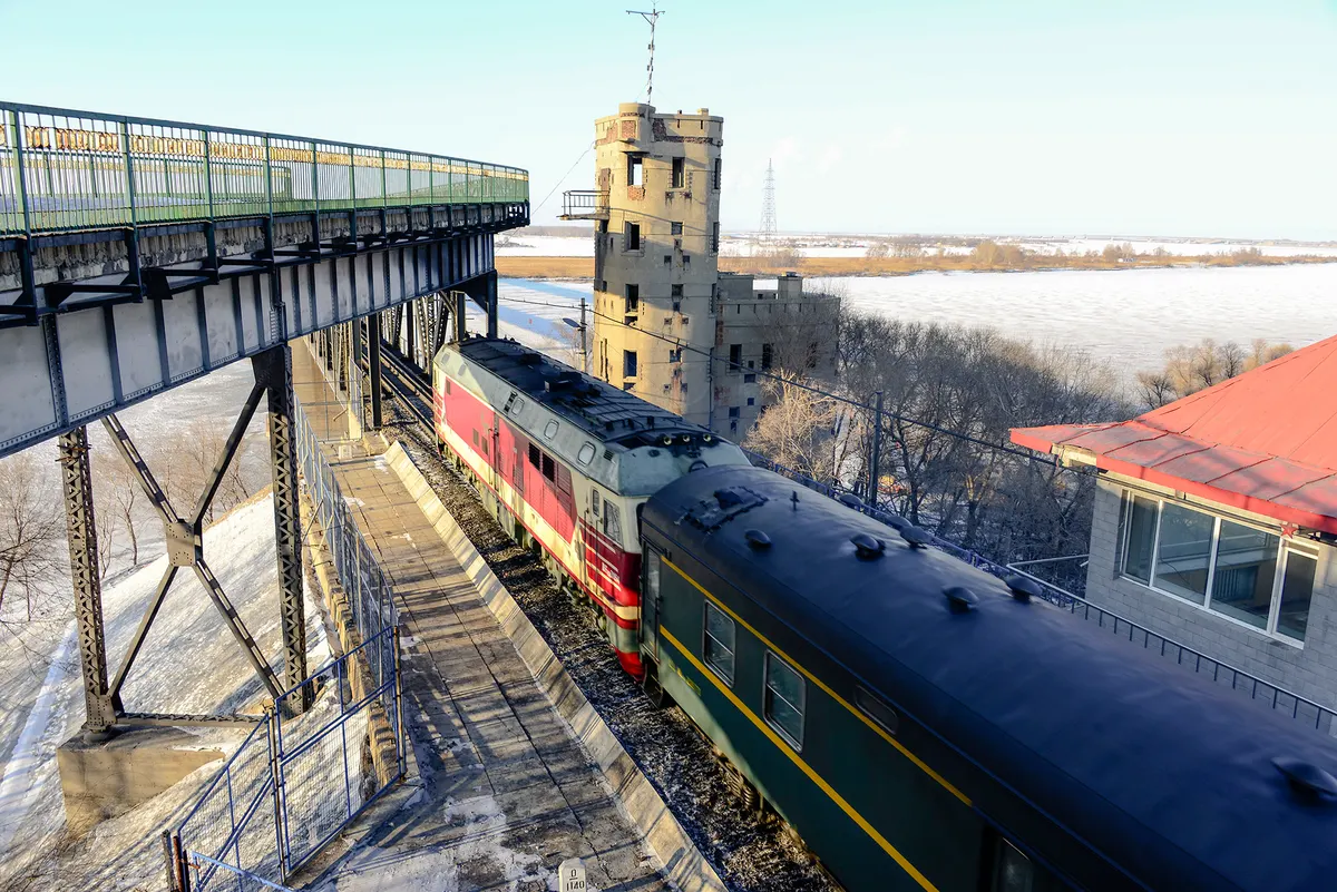 A diesel locomotive pulling green carriages off an old steel railway bridge beside a stone watchtower, with the frozen river and snowy bank beyond