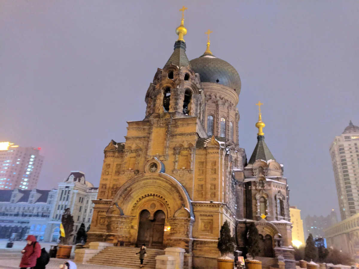 St Sophia Cathedral floodlit in gold at dusk, with snow on its ledges and domes, a few people on the square and tower blocks behind
