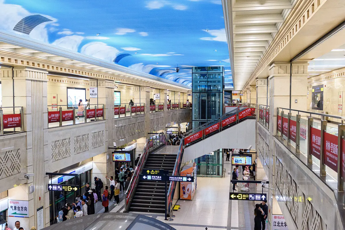 A two-level metro station hall under a ceiling painted as a blue sky with clouds, with a staircase, a glass lift and passengers on both levels