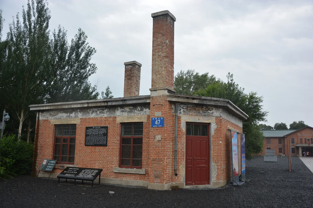 A small single-storey red-brick building with two tall brick chimneys and an information board in front, on a gravel site with trees behind