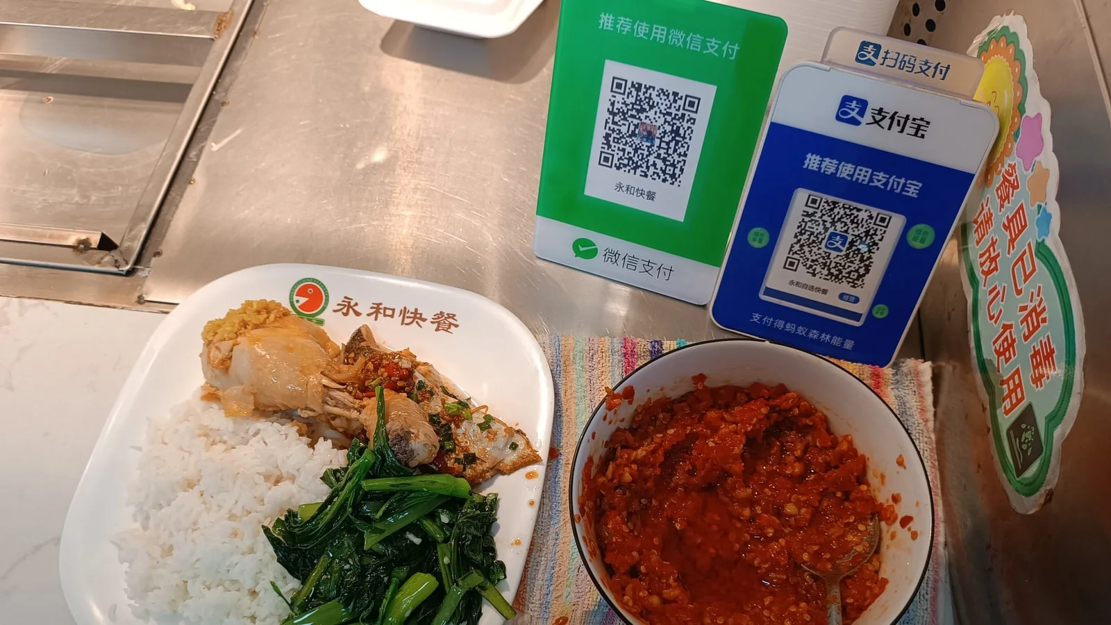 A canteen lunch of rice, chicken and greens on a counter beside green WeChat Pay and blue Alipay QR code stands
