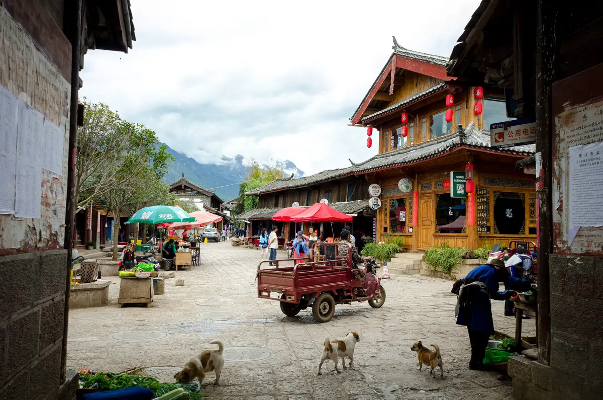 A village street in Baisha with timber houses hung with red lanterns, market stalls under umbrellas, a three-wheeled cart and dogs crossing the paving