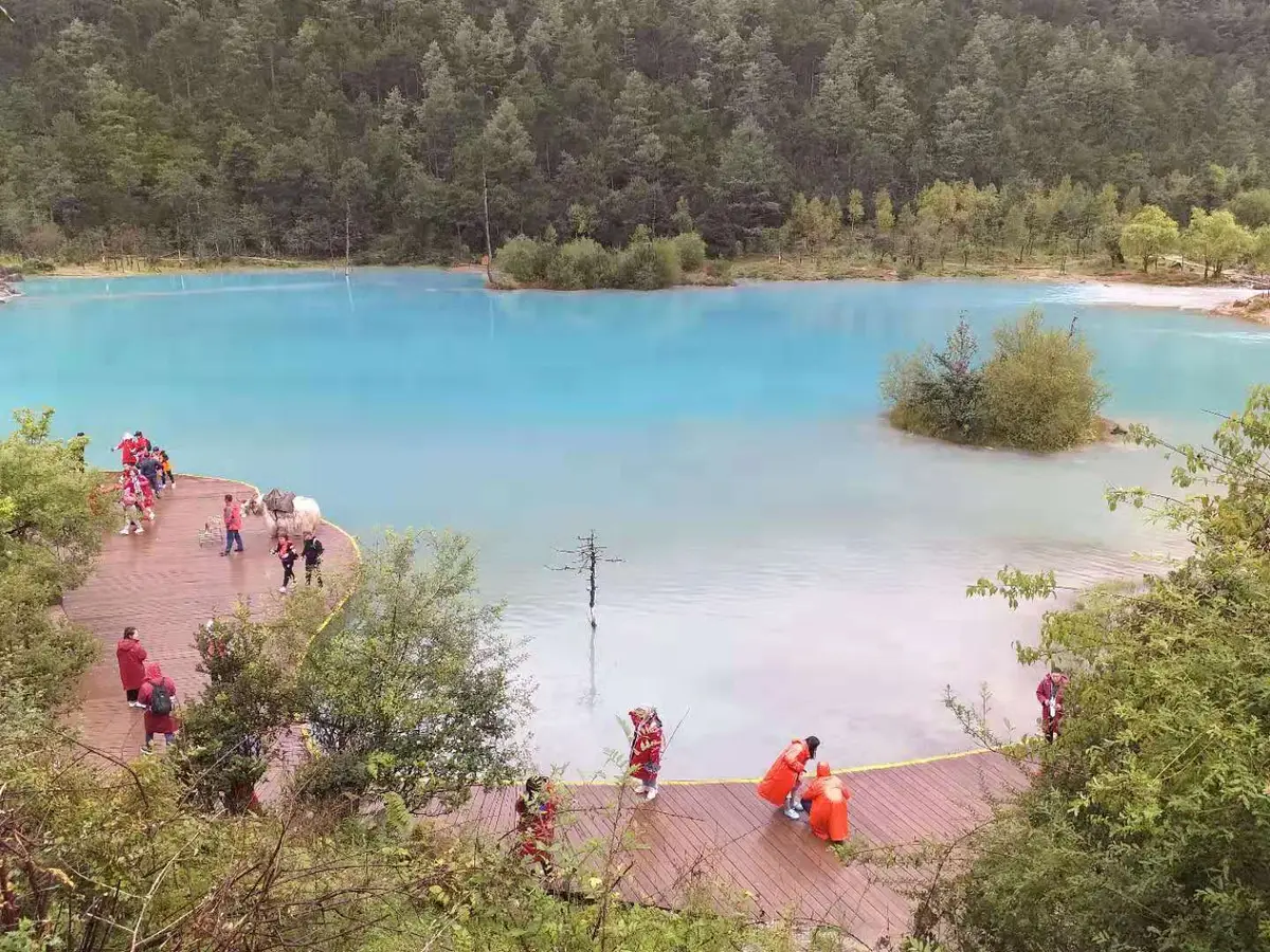 A pool of opaque turquoise water ringed by forest, with a wooden boardwalk along its edge and visitors in red jackets