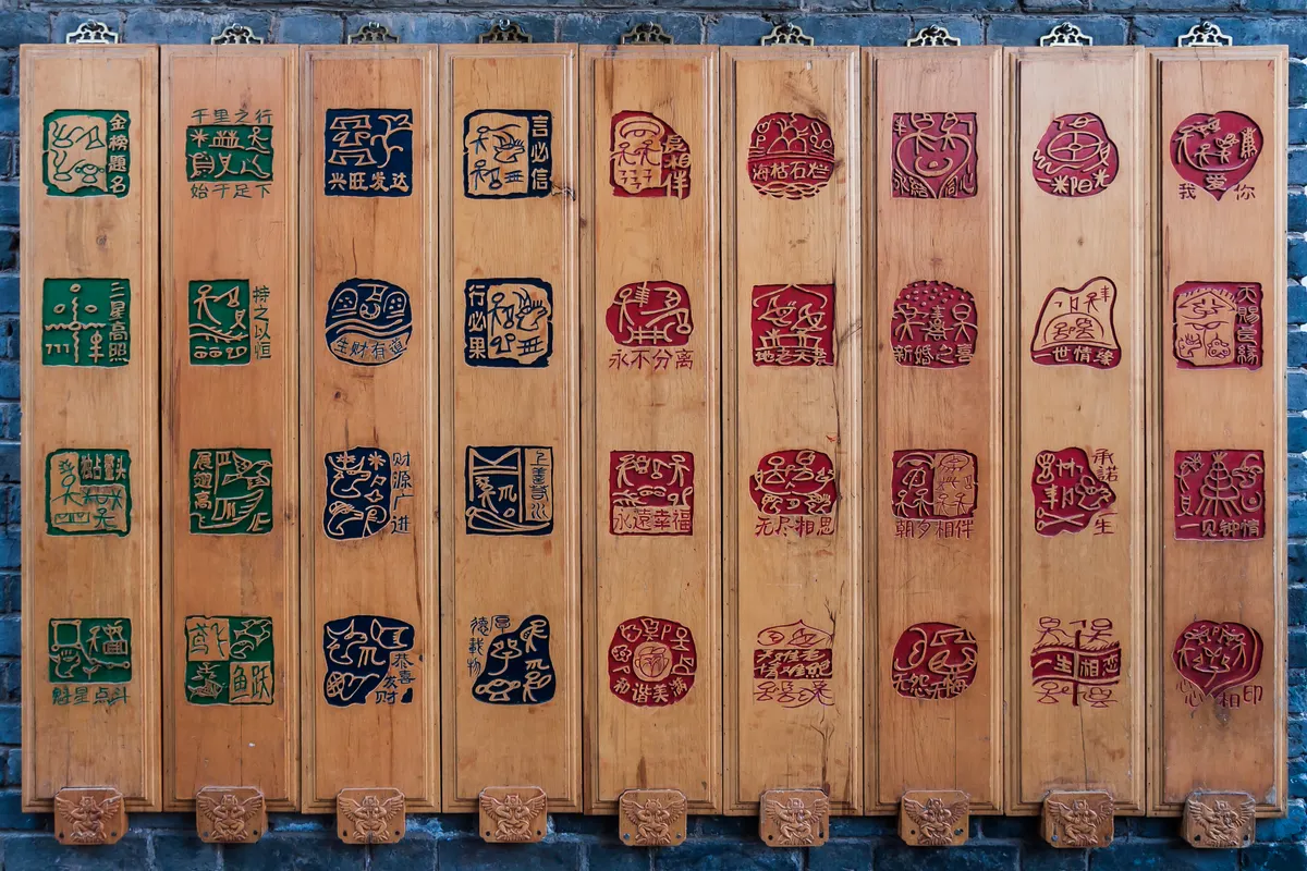 Nine tall wooden boards hung on a grey brick wall, each carved with four Dongba pictograph seals in green, dark blue or red, with Chinese characters beside them