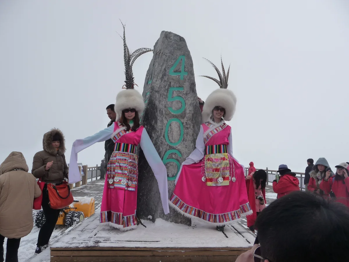 Two women in pink Naxi costumes and white fur hats posing beside a tall stone engraved with the number 4506, on a snowy viewing platform with other visitors in warm coats