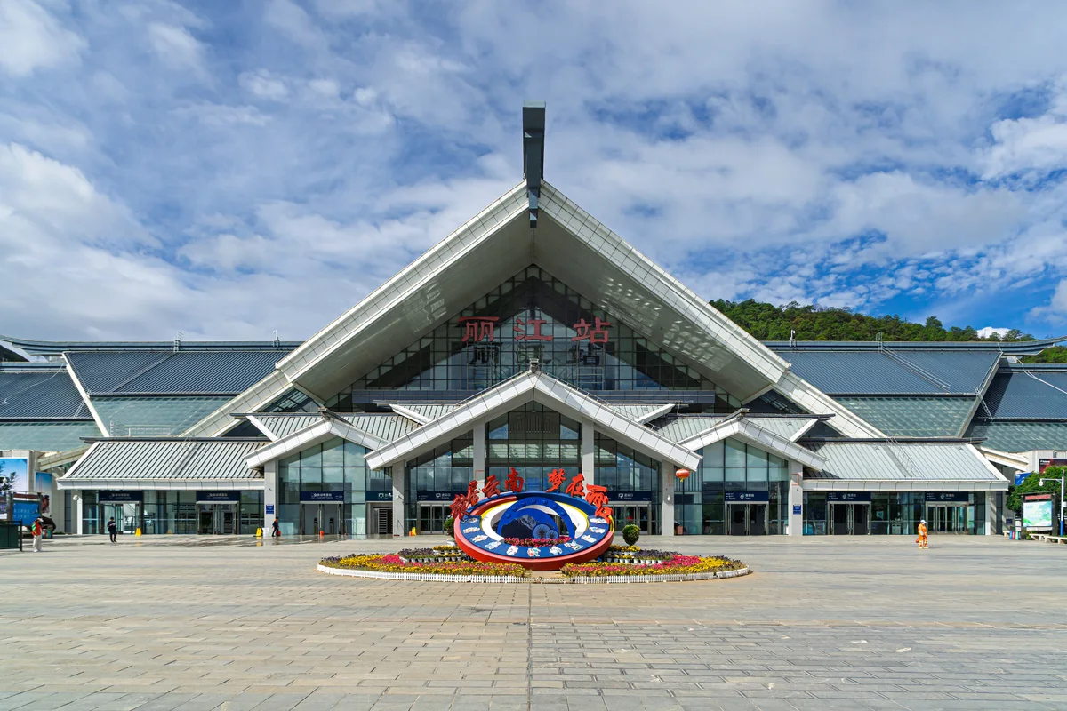 The front of Lijiang railway station, a long glass hall under a steep pointed roof, with a flower bed and a wide empty forecourt under a cloudy blue sky