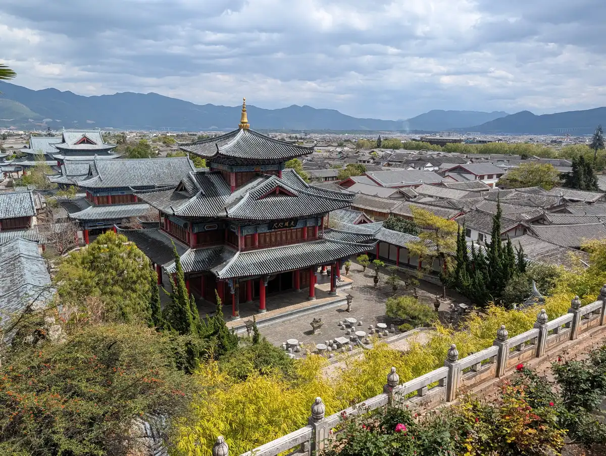 A red and dark-roofed tiered pavilion inside a walled compound, with the grey roofs of the Old Town and a range of mountains beyond