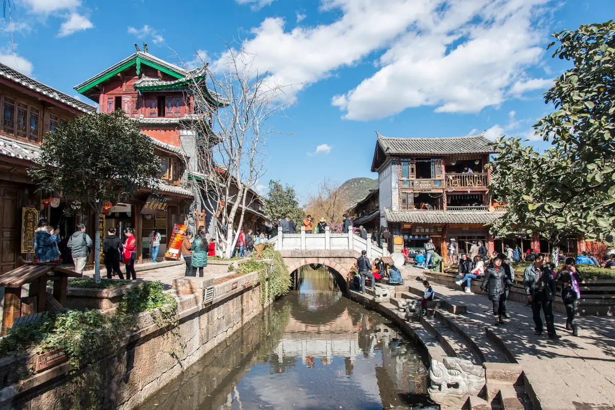 A stone-banked canal running between two-storey timber houses in Lijiang Old Town, with a small arched bridge and visitors on the paved quay under a blue sky