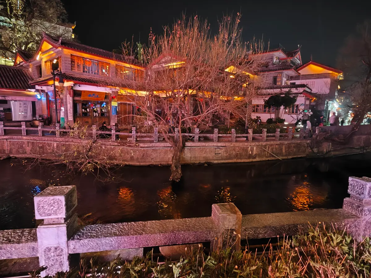 Timber buildings lit in warm orange and pink along a canal at night in Lijiang Old Town, with a stone balustrade in the foreground