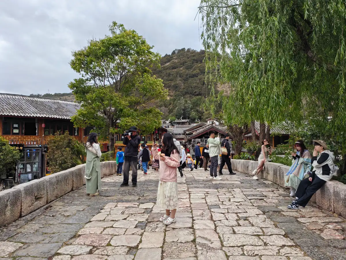 A broad stone bridge paved with worn slabs in Shuhe, with visitors posing for photos, willow trees and timber houses behind