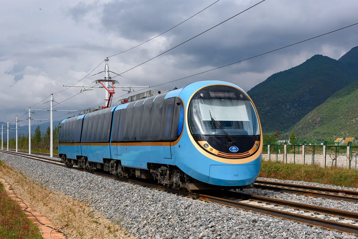 A sky-blue articulated tram-train on a ballasted track under overhead wires, with green mountains and grey cloud behind