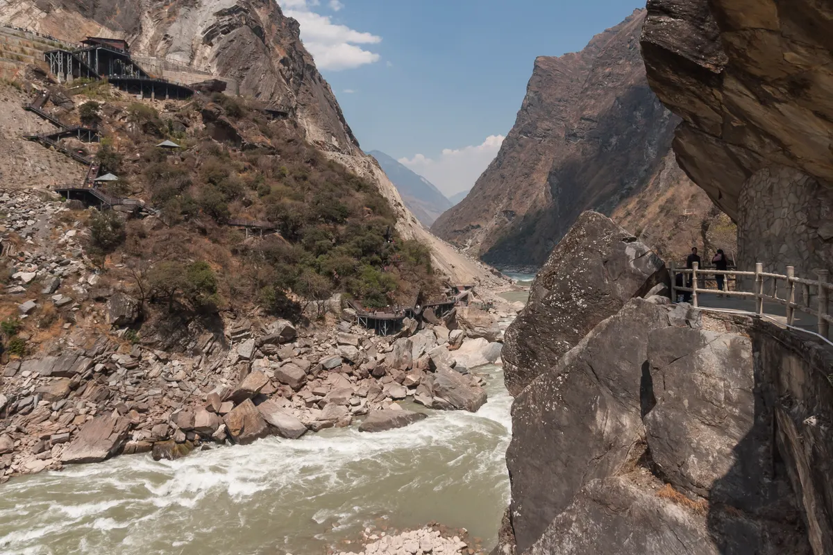 The Jinsha River rushing over boulders at the bottom of a deep rocky gorge, with a railed path cut into the cliff on the right and steep mountainsides above