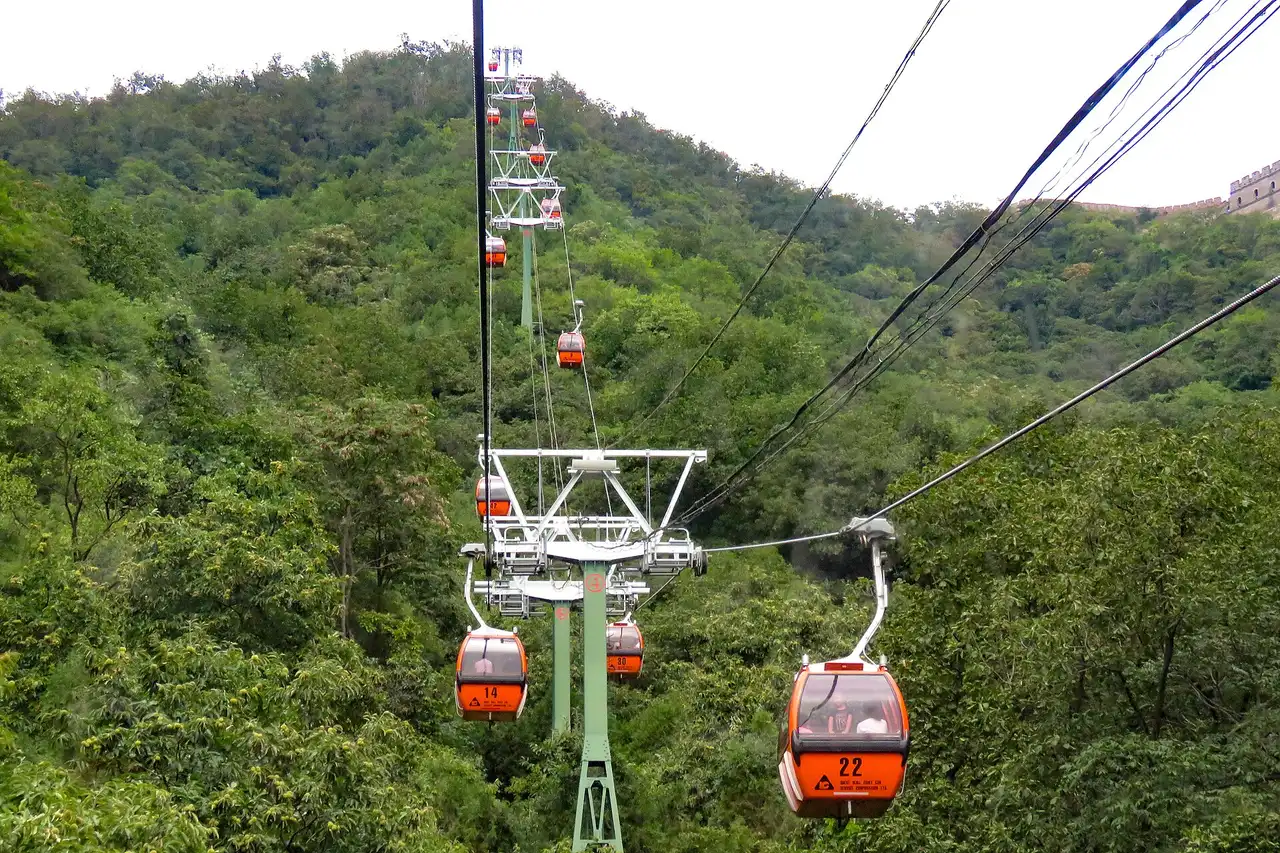 Orange gondolas of the Mutianyu cable car climbing a forested hillside, with the Great Wall on the ridge at top right