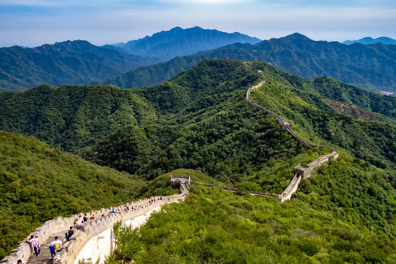 The Great Wall at Mutianyu winding along green forested ridges, with watchtowers on the crest, visitors on the near stretch and mountains behind