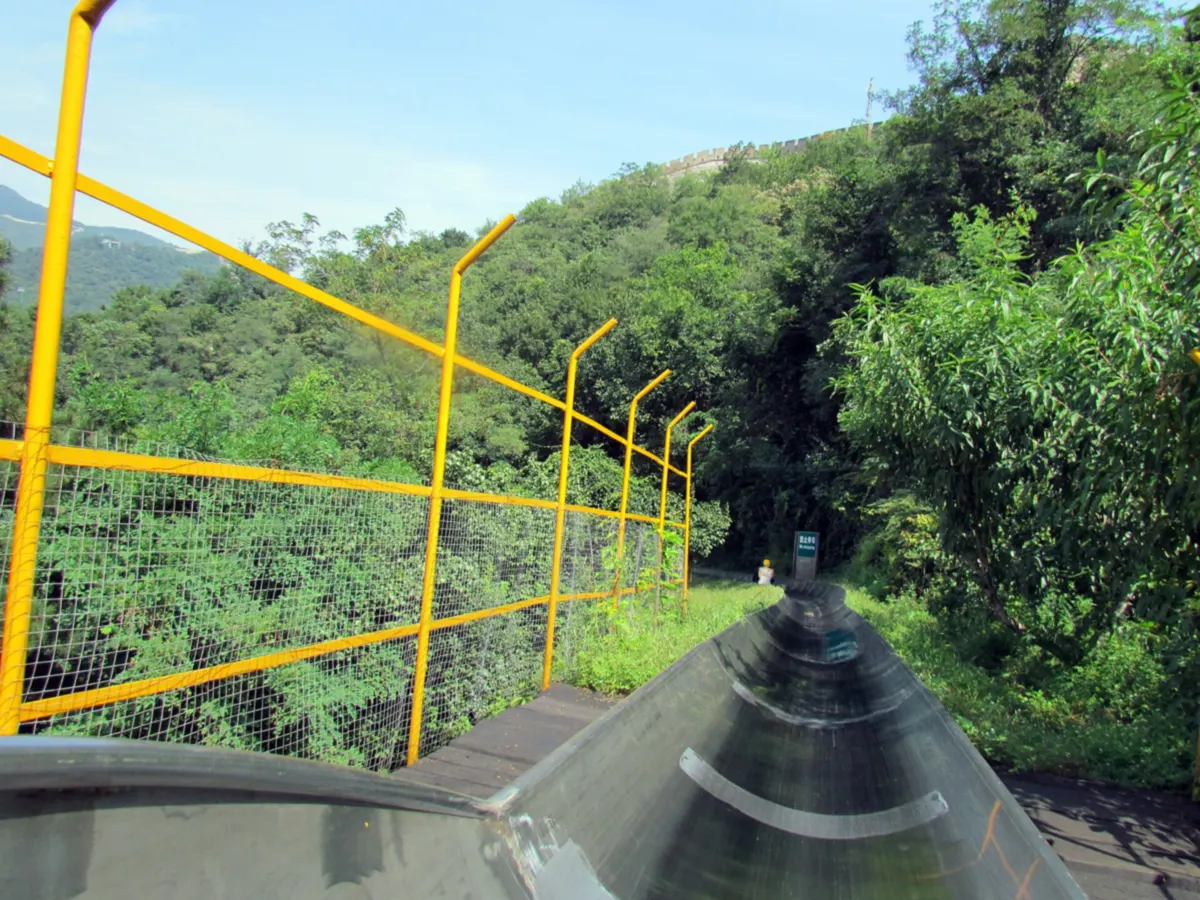 The metal chute of the Mutianyu toboggan running down through trees, with the wall on the hill above
