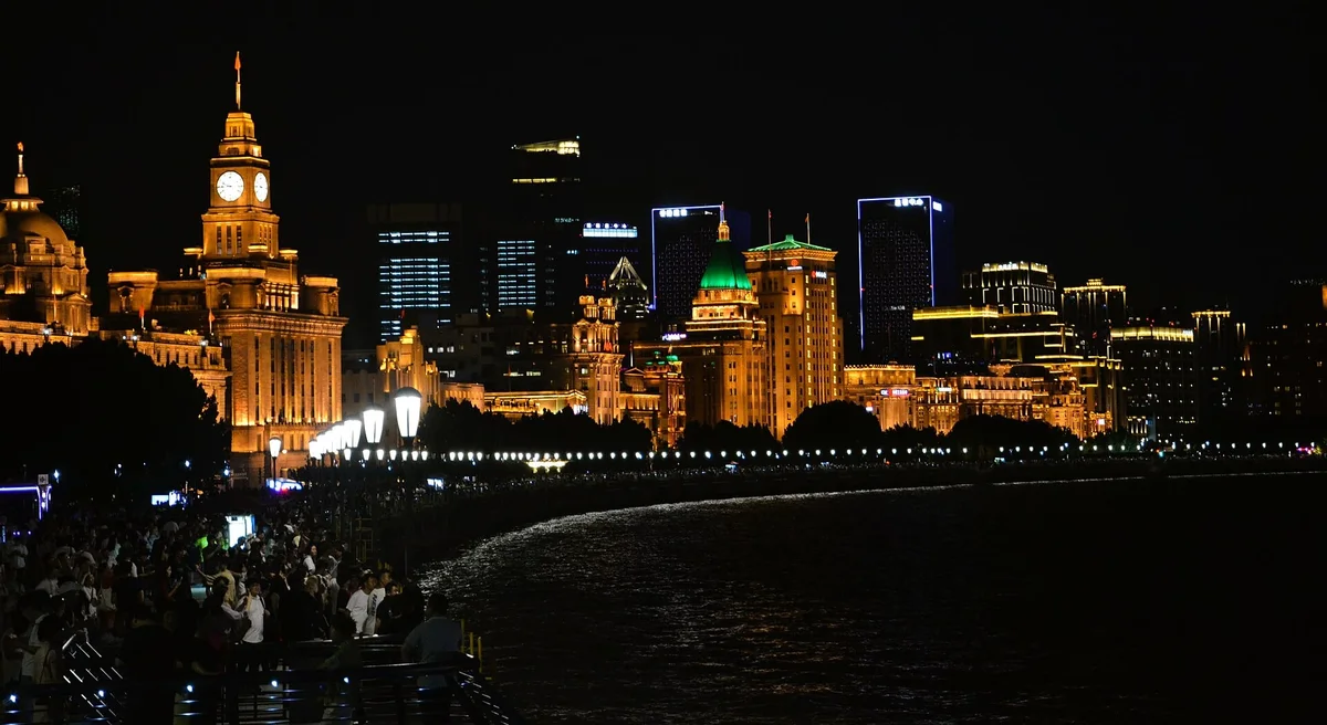 Crowds along the Bund promenade at night beside the dark river, with the lit clock tower of the Customs House and old bank buildings glowing gold