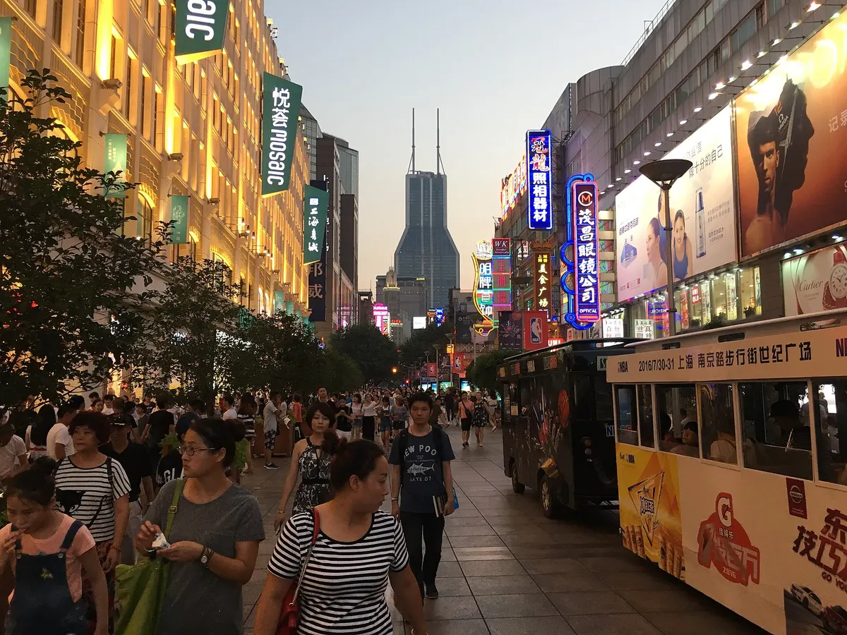 Evening crowds walking along the pedestrian stretch of East Nanjing Road under neon shop signs, with a small sightseeing tram on the right