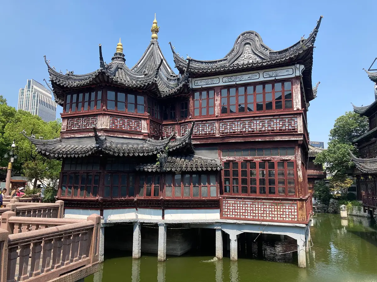 A two-storey wooden teahouse with upswept roofs standing on stilts in a green pond, with stone bridge railings in front