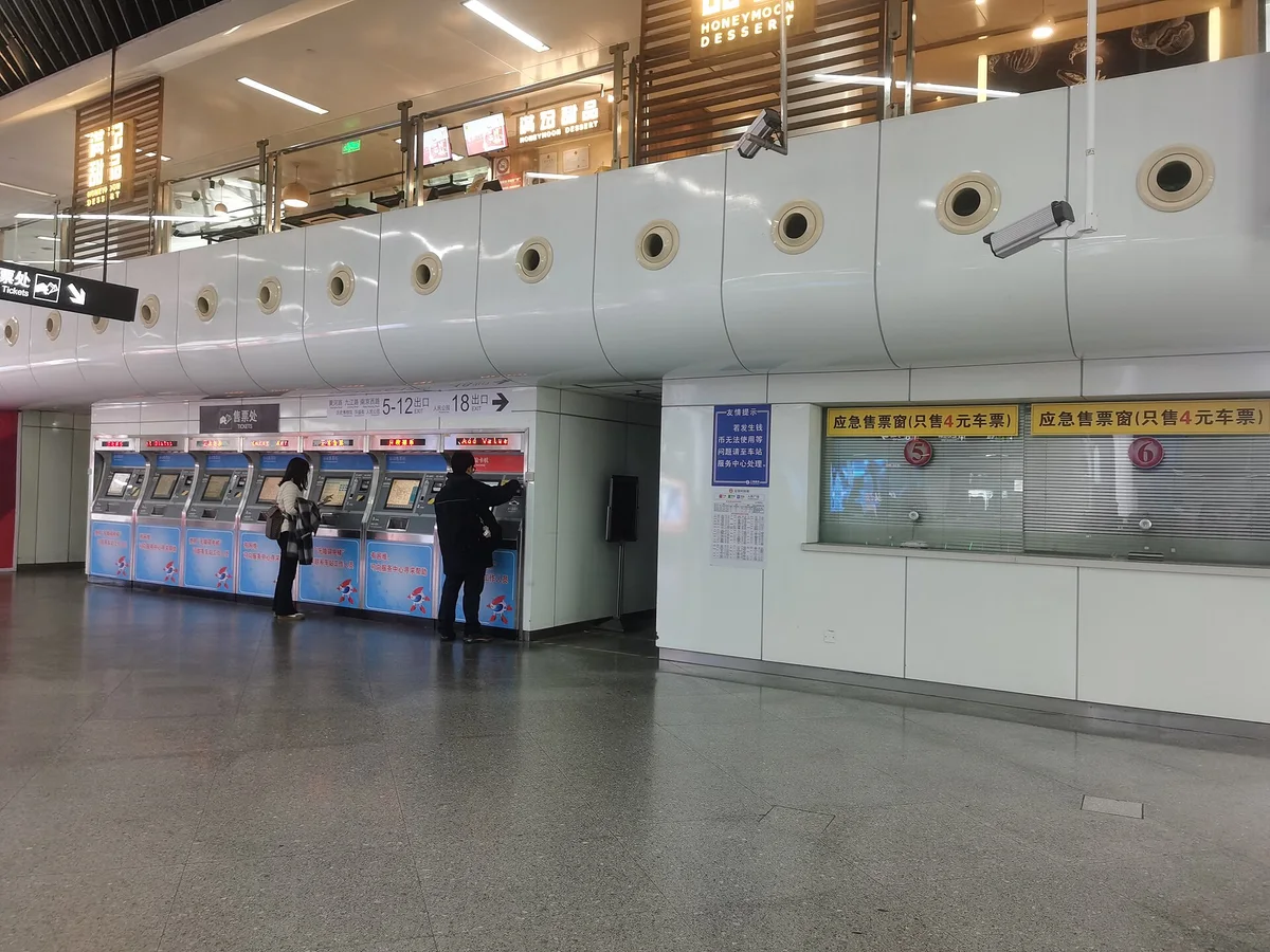 A row of blue ticket vending machines, two passengers buying tickets, and two emergency ticket windows with their blinds down in a bright metro station concourse