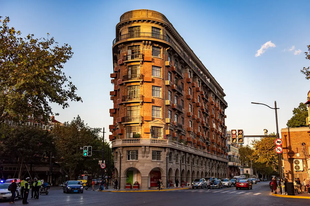 The wedge-shaped red-brick Wukang Building with rows of balconies on a street corner in late afternoon sun, plane trees and traffic around it
