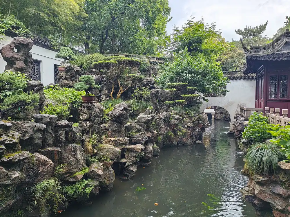 Grey rockeries planted with pines and shrubs beside a narrow stream, with a white wall and a pavilion with upturned eaves in Yu Garden