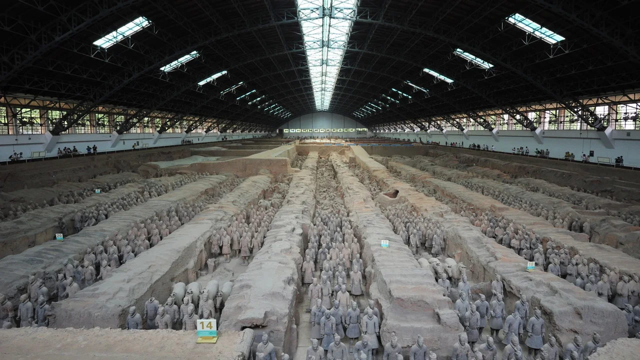 Rows of terracotta warriors standing in the earth corridors of Pit 1 under the arched steel roof of its hall, with visitors along the far railing