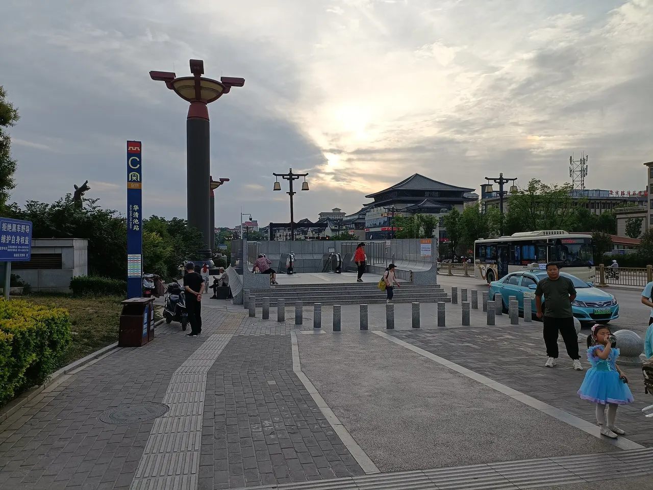 A metro exit with escalators under a grey sky, a tall blue sign marked C for Huaqingchi station, a taxi and a bus at the kerb