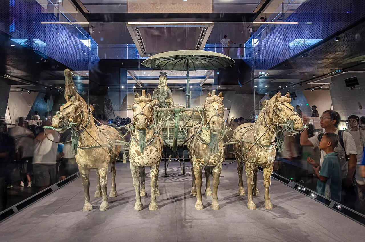 A half-size bronze chariot with a parasol canopy, drawn by four bronze horses, in a glass case with visitors behind it