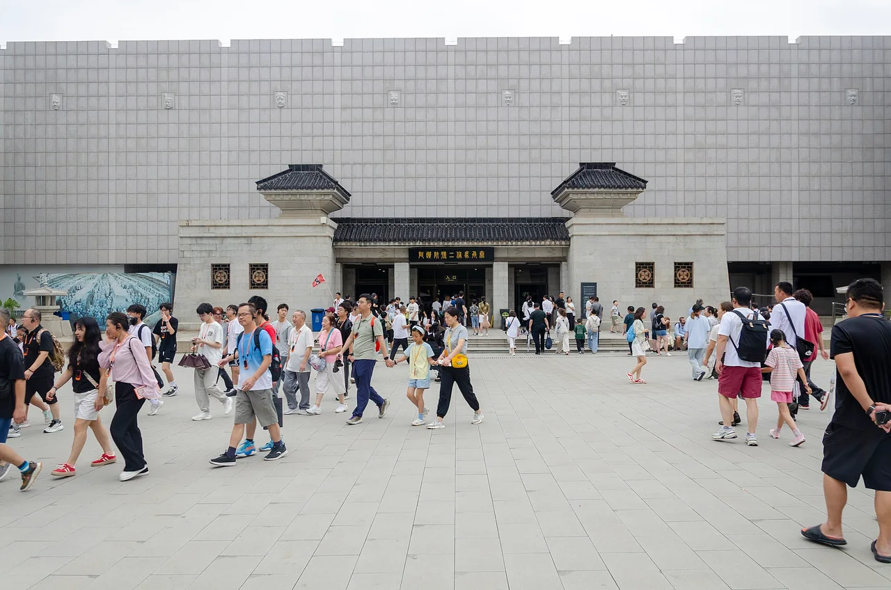 A crowd of visitors, many wearing lanyards and earpieces, crossing a paved square in front of the grey stone entrance to the Pit 2 hall
