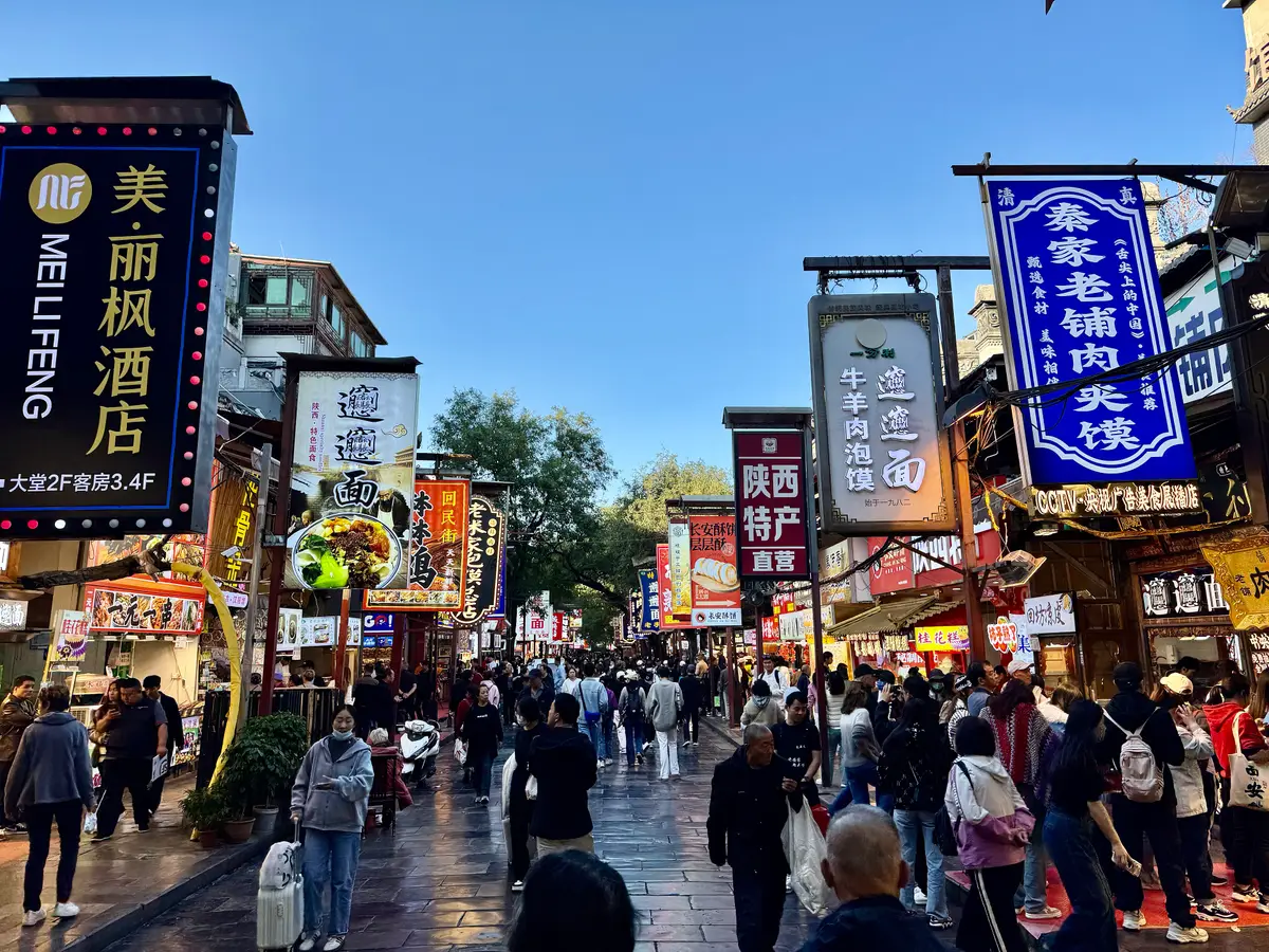 A crowded pedestrian street hung with shop signs in Chinese characters, with food stalls on both sides under a blue sky