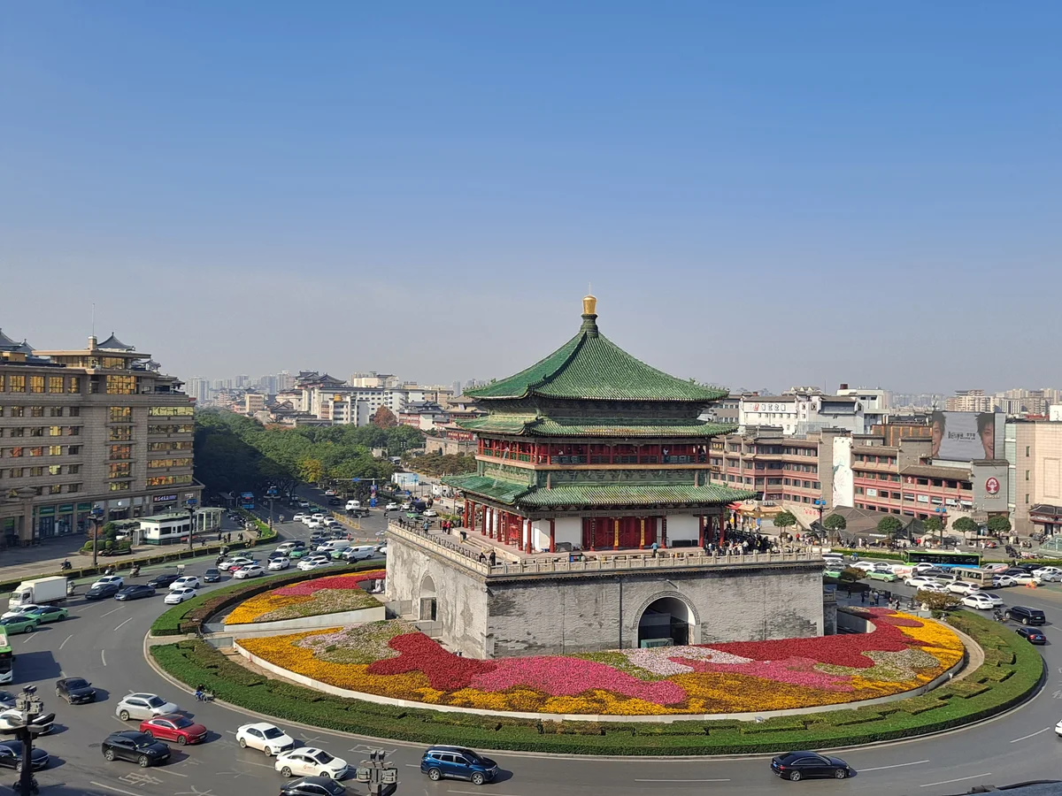 The Bell Tower with its green-tiled triple roof on a stone base, surrounded by flower beds on a traffic island with cars circling it