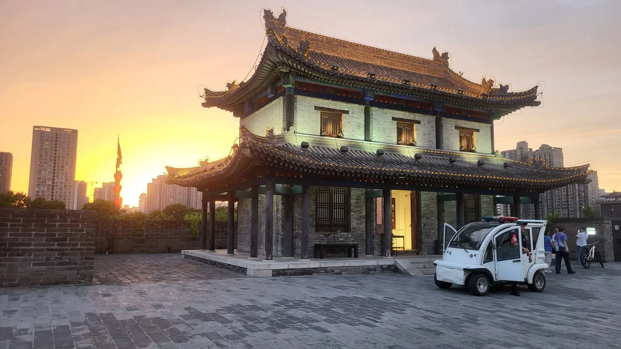 A two-storey watchtower pavilion on top of the Xi'an city wall at sunset, with a white patrol cart on the paved walkway and tower blocks beyond the parapet