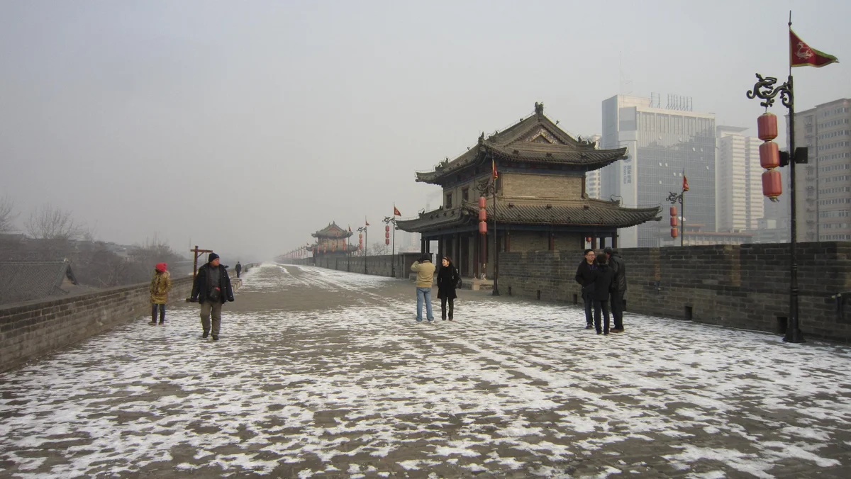 Patchy snow on the paved top of the city wall, with a few people walking towards a gate tower and grey high-rises behind