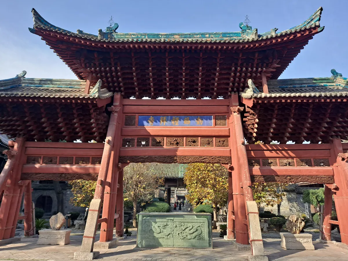 A red wooden archway with a heavy tiled roof and carved brackets in a mosque courtyard, with a stone slab carved with dragons and two decorative rocks on plinths beneath it