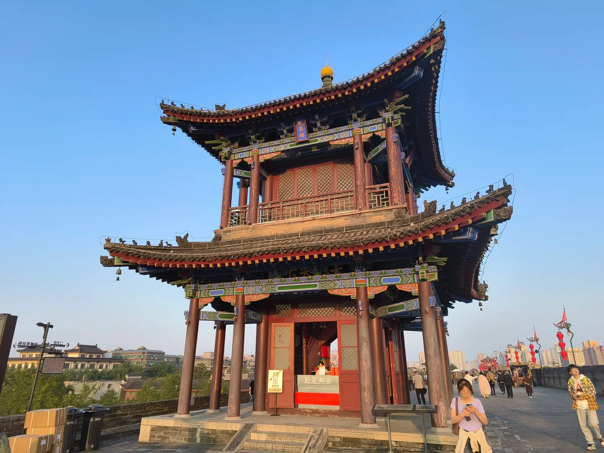 A two-tiered red and green pavilion with upturned eaves on top of the city wall, with visitors walking past in late sun