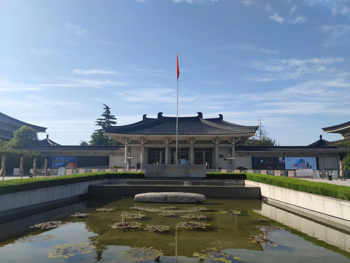 A low building with a wide Tang-style roof behind a rectangular pond with water lilies and a flagpole, under a blue sky