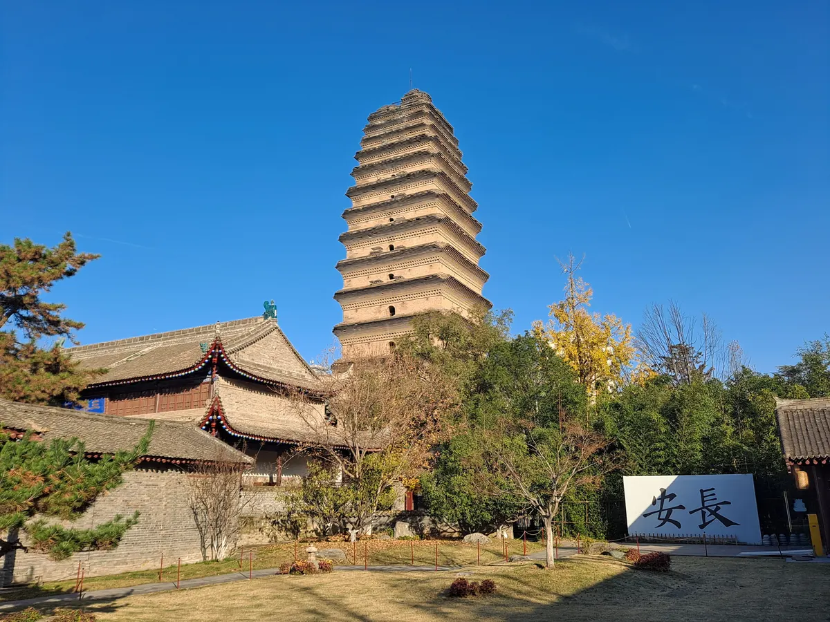 A slender many-tiered brick pagoda above the grey tiled roofs of temple halls, with pine and yellow ginkgo trees under a clear sky