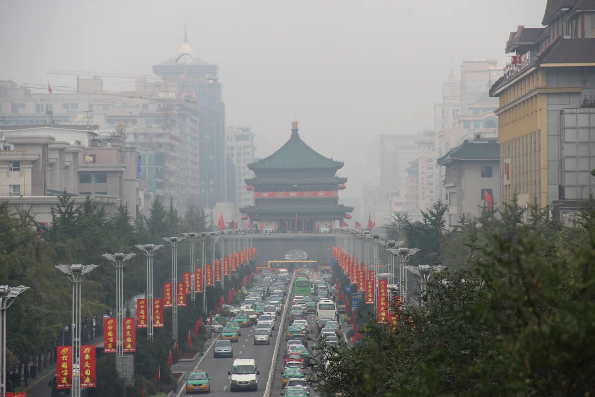 A wide avenue lined with red banners and lamp posts leading to the green-roofed Bell Tower, seen from above through thick grey smog