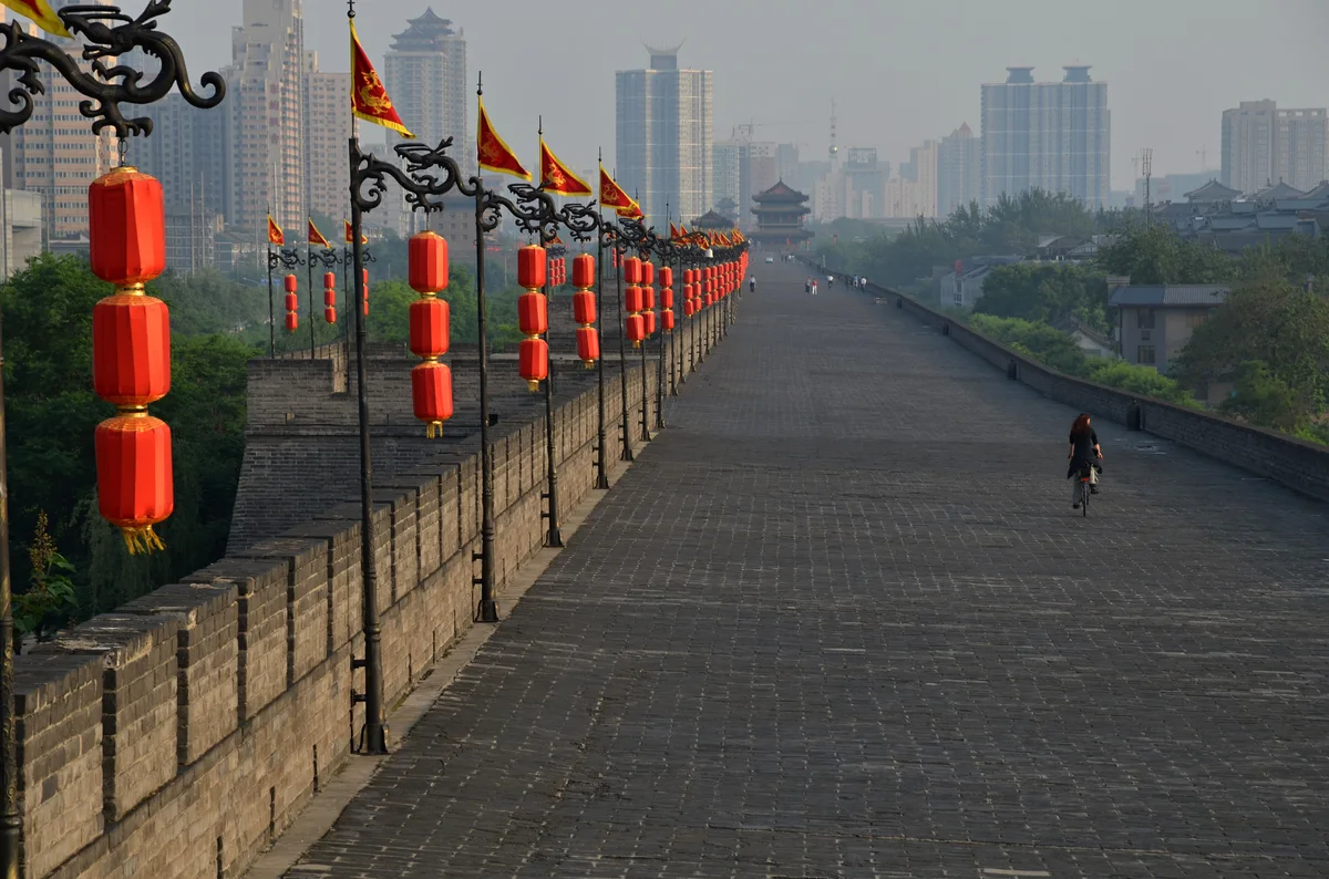 A cyclist riding along the wide paved top of the city wall beside a row of red lanterns, with high-rise towers beyond
