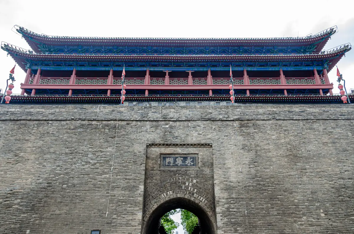 The grey brick face of the South Gate with a stone name plaque above its arched passage and a long red and blue gate tower on top